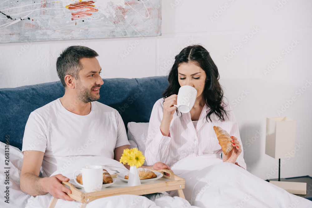 happy man looking at attractive woman drinking coffee and holding croissant in bed happy man looking at attractive woman drinking coffee and holding croissant in bed