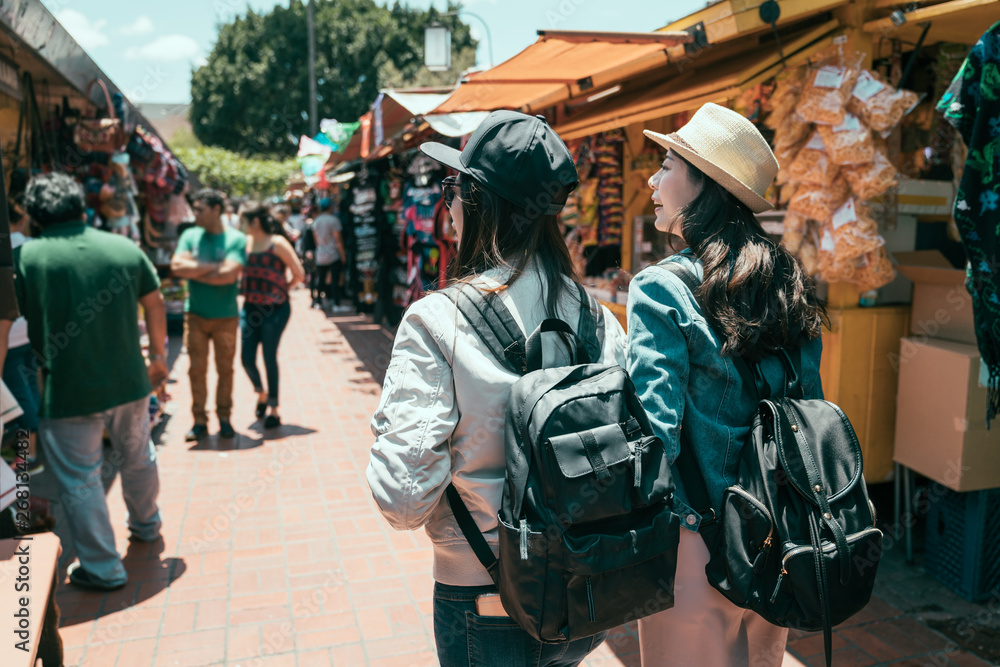back view of two asian girls best friends travel in los angeles together on summer break. young women tourist backpacker shopping in mexico outdoor market. ladies walking in olvera street having fun.
