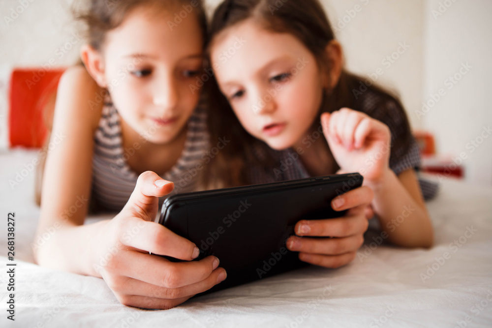 Children using digital gadgets at home. Little girls playing games on the tablet lying on the bed in the bedroom.