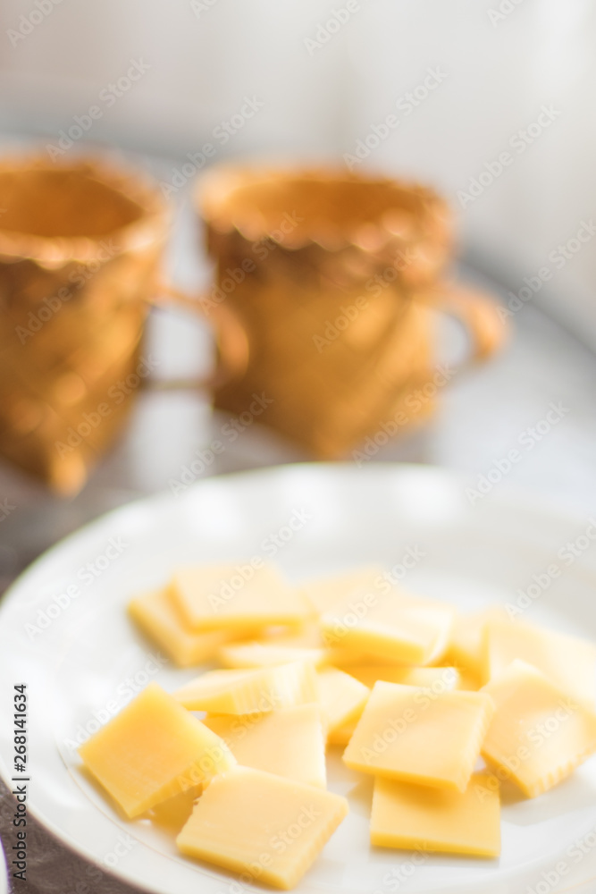 Semi-hard cheese sliced in white plate. Two wicker cups on a sunny table Semi-hard cheese sliced in white plate. Two wicker cups on a sunny table