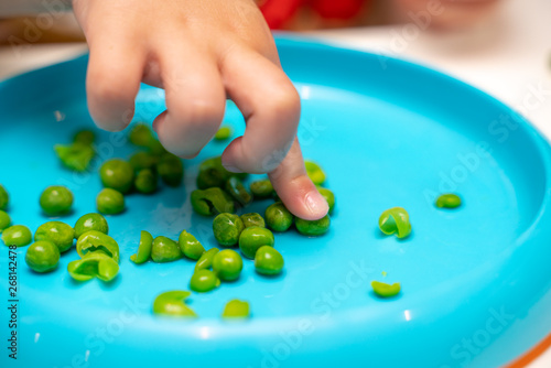Young child s hands picking up  playing with and eating peas at dinner time from a blue plate