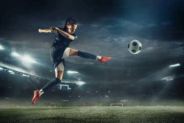 Young female soccer or football player with long hair in sportwear and boots kicking ball for the goal in jump at the stadium. Concept of healthy lifestyle, professional sport, hobby, motion, movement