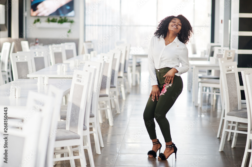 Obraz premium Сheerful business african american lady with afro hair, wear white blouse and green pants posed in cafe.