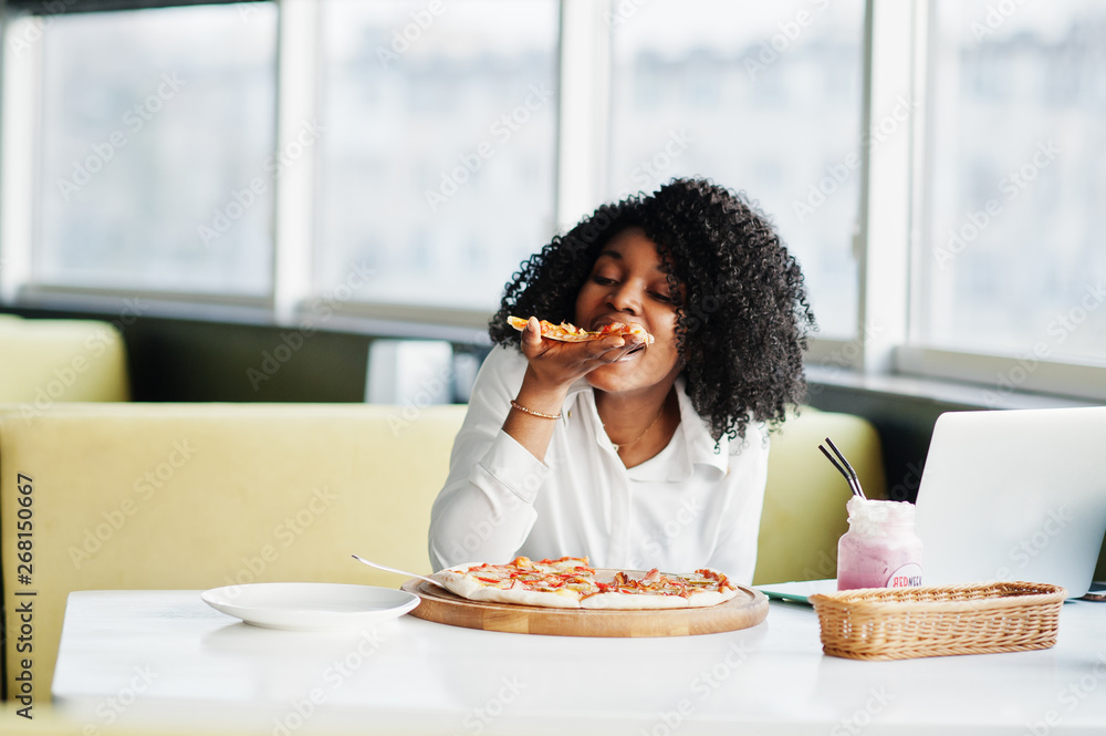 Сheerful business african american lady with afro hair, wear white blouse sitting at table, work with laptop in cafe, eat pizza and drink pink milkshake cocktail. Сheerful business african american lady with afro hair, wear white blouse sitting at table, work with laptop in cafe, eat pizza and drink pink milkshake cocktail.