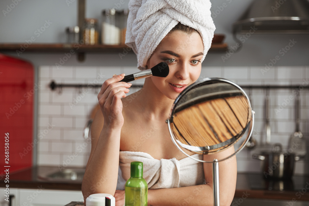 Photo of satisfied caucasian woman looking in mirror and applying makeup with cosmetic brush in apartment