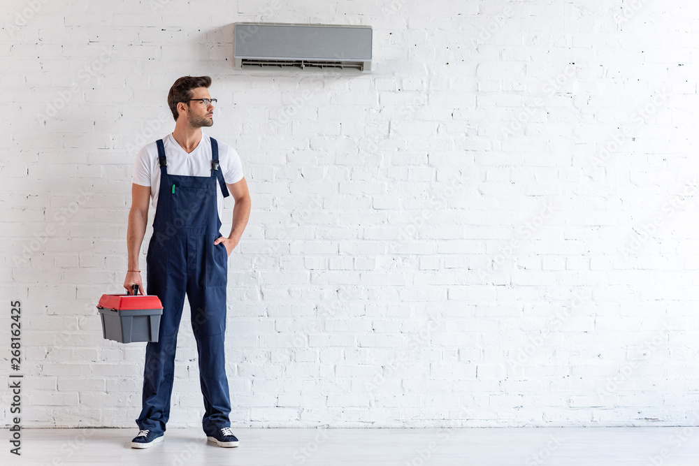 handsome repairman in uniform standing under conditioner and looking away handsome repairman in uniform standing under conditioner and looking away