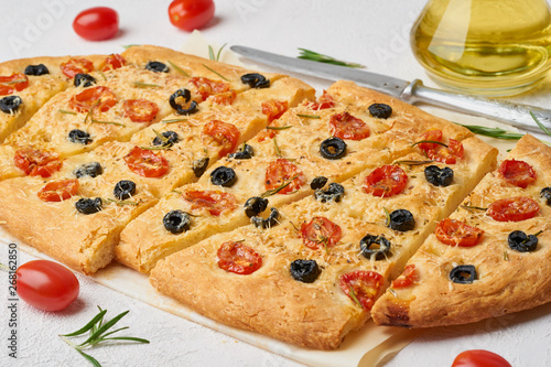 Focaccia, pizza with tomatoes, olives and rosemary. Chopped Italian flat bread. Side view, white concrete background