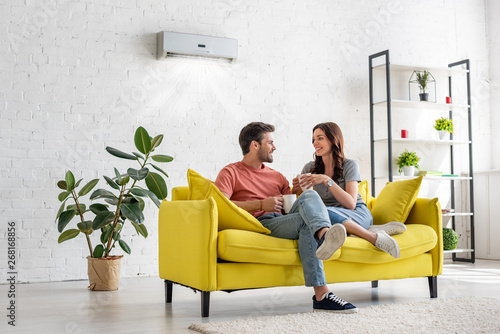 happy young couple talking and holding cups while sitting on yellow sofa under air conditioner at home photo