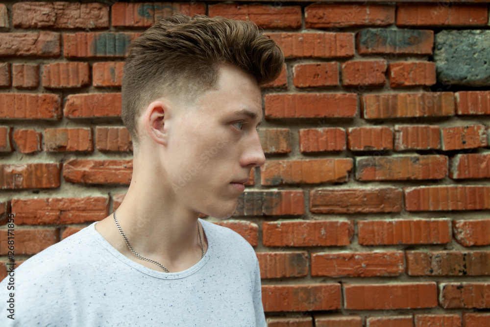 young blond guy with a fashionable haircut in a white t-shirt in profile against a brick wall