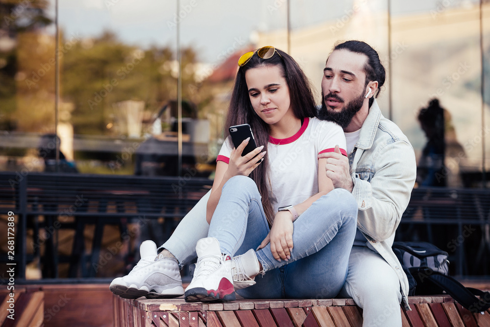 Naklejka premium Beautiful young happy couple boyfriend and girlfriend resting together during a romantic trip to a European country and watching a video about the sights using a smartphone and wireless internet