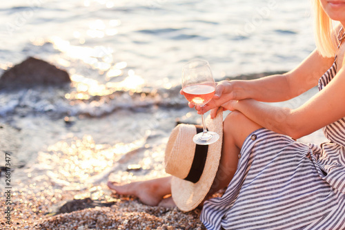 Woman is sitting on sea beach with wineglass of pink wine at sunset in summer vacation in resort. Tourist girl in striped dress with straw hat is enjoying life, relaxing, drinking, traveling.