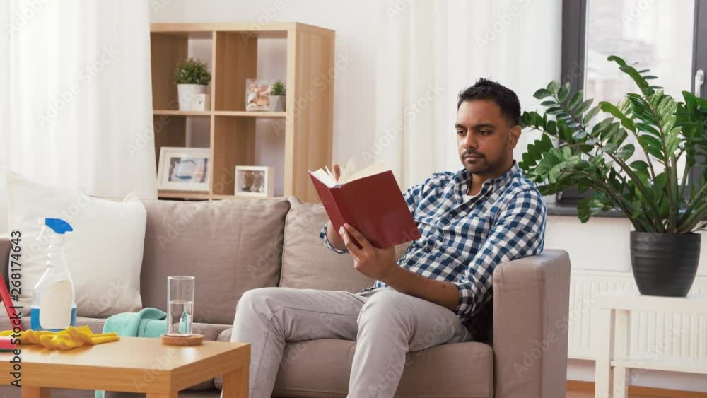 household and people concept - indian man reading book and resting after home cleaning and drinking water from glass