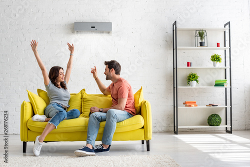 happy man and woman talking while sitting on yellow sofa under air conditioner at home