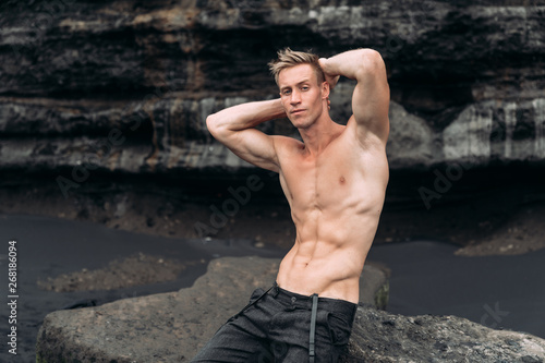 Portrait of young man with naked torso siting on rocky beach with black sand.