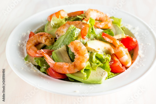 Fresh shrimp salad made of tomato, ruccola, avocado, prawns, chicken breast, arugula, crackers and spices. Caesar salad in a white, transparent bowl on wooden background