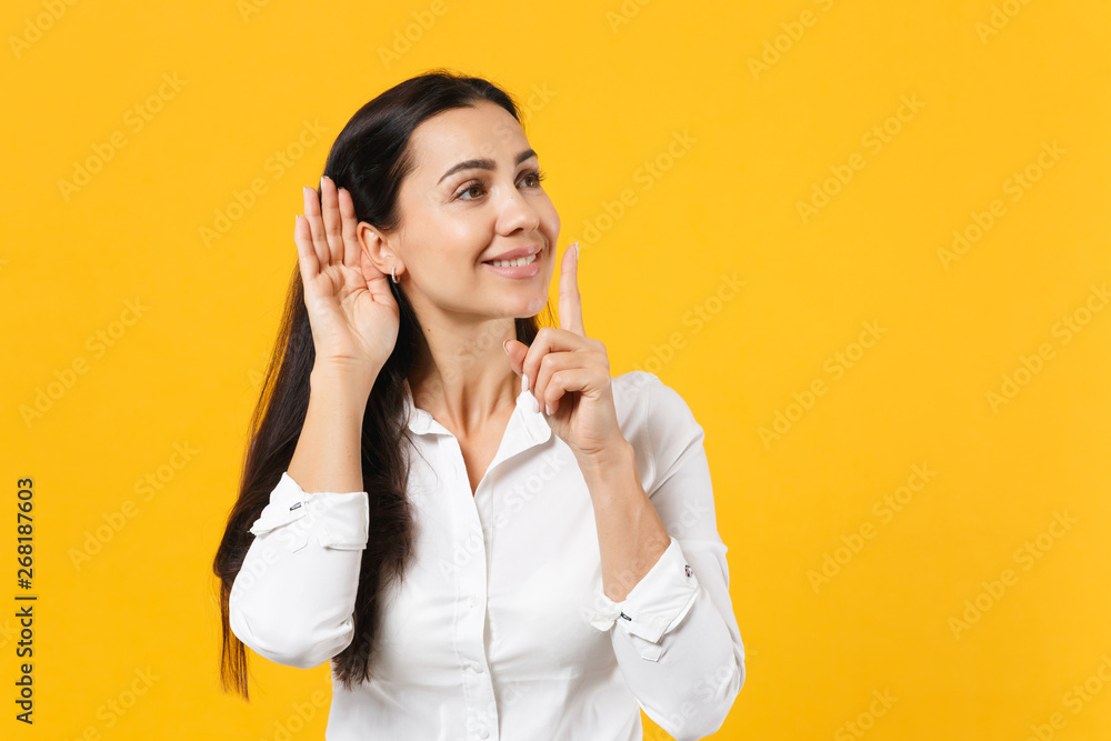 Portrait of smiling young woman in white shirt saying hush be quiet with finger on lips shhh gesture try to hear you isolated on yellow orange background. People lifestyle concept. Mock up copy space. Portrait of smiling young woman in white shirt saying hush be quiet with finger on lips shhh gesture try to hear you isolated on yellow orange background. People lifestyle concept. Mock up copy space.