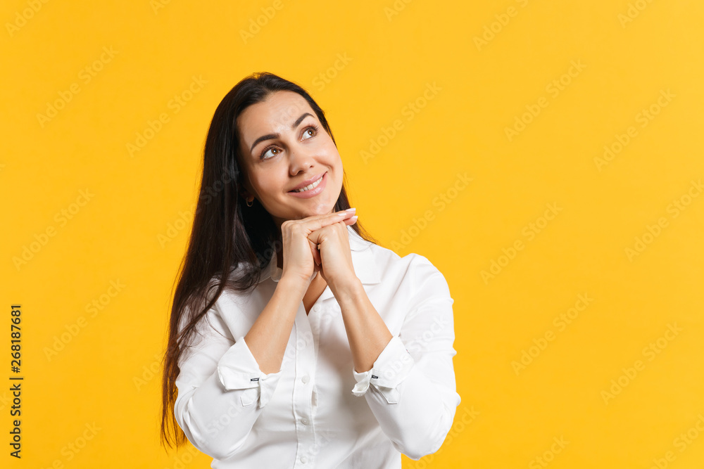 Portrait of pensive smiling young woman in white shirt put hand prop up on chin, looking up isolated on bright yellow orange wall background in studio. People lifestyle concept. Mock up copy space. Portrait of pensive smiling young woman in white shirt put hand prop up on chin, looking up isolated on bright yellow orange wall background in studio. People lifestyle concept. Mock up copy space.