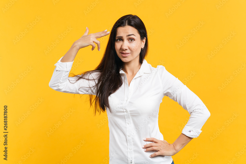 Portrait of desperate young woman in white casual shirt point finger to head as if she about shoot herself isolated on yellow orange background in studio. People lifestyle concept. Mock up copy space. Portrait of desperate young woman in white casual shirt point finger to head as if she about shoot herself isolated on yellow orange background in studio. People lifestyle concept. Mock up copy space.