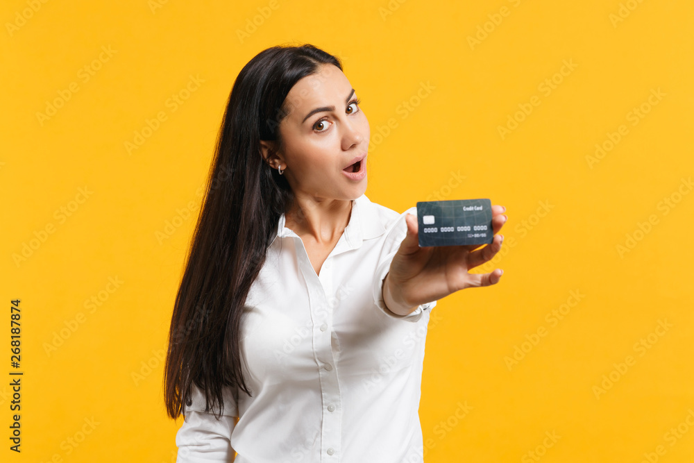 Portrait of shocked young woman in white casual shirt holding credit bank card, keeping mouth open isolated on yellow orange wall background in studio. People lifestyle concept. Mock up copy space. Portrait of shocked young woman in white casual shirt holding credit bank card, keeping mouth open isolated on yellow orange wall background in studio. People lifestyle concept. Mock up copy space.