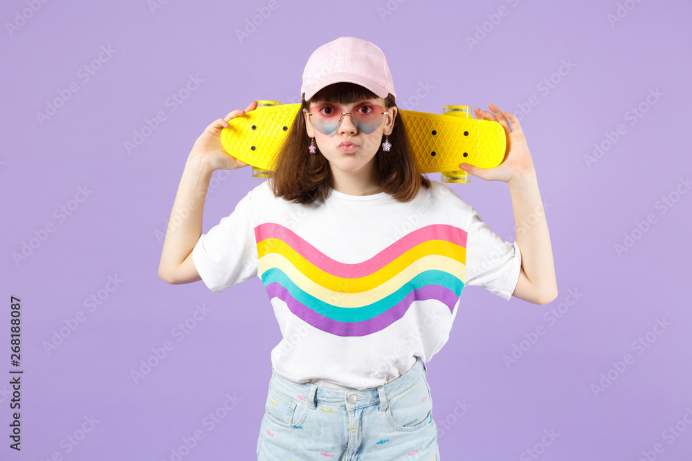 Pretty teen girl in vivid clothes, heart eyeglasses holding yellow skateboard, blowing lips isolated on violet pastel wall background. People sincere emotions, lifestyle concept. Mock up copy space.