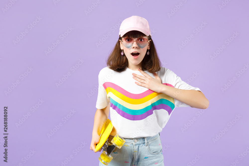 Amazed teen girl in vivid clothes, eyeglasses with yellow skateboard keeping mouth open holding hand on chest isolated on violet pastel background. People sincere emotions concept. Mock up copy space. Amazed teen girl in vivid clothes, eyeglasses with yellow skateboard keeping mouth open holding hand on chest isolated on violet pastel background. People sincere emotions concept. Mock up copy space.