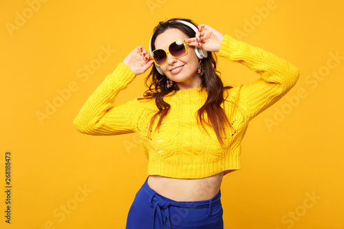 Attractive woman in sweater, blue trousers, sunglasses listen music with headphones looking aside isolated on yellow orange background. People sincere emotions, lifestyle concept. Mock up copy space.