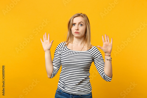 Portrait of perplexed young woman in striped clothes rising hands, showing palms isolated on yellow orange wall background in studio. People sincere emotions lifestyle concept. Mock up copy space.