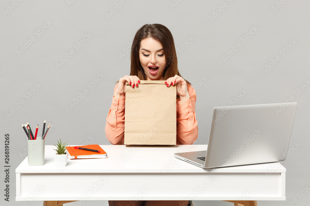 Young amazed woman in pastel clothes holding brown clear empty blank craft paper bag for takeaway work at desk with laptop isolated on gray background. Achievement business career lifestyle concept. Young amazed woman in pastel clothes holding brown clear empty blank craft paper bag for takeaway work at desk with laptop isolated on gray background. Achievement business career lifestyle concept.