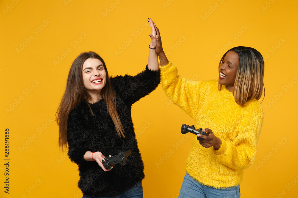 Two fun young women friends european and african american in black yellow clothes hold joystick isolated on bright orange wall background, studio portrait. People lifestyle concept. Mock up copy space Two fun young women friends european and african american in black yellow clothes hold joystick isolated on bright orange wall background, studio portrait. People lifestyle concept. Mock up copy space
