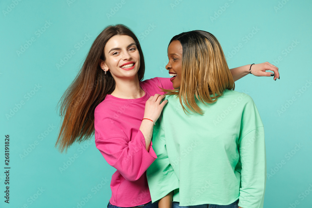 Two young women friends european and african american in pink green clothes standing posing isolated on blue turquoise wall background, studio portrait. People lifestyle concept. Mock up copy space. Two young women friends european and african american in pink green clothes standing posing isolated on blue turquoise wall background, studio portrait. People lifestyle concept. Mock up copy space.