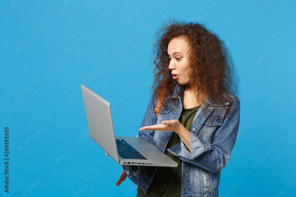 Young african american girl teen student in denim clothes, backpack work on pc isolated on blue wall background studio portrait. Education in high school university college concept. Mock up copy space Young african american girl teen student in denim clothes, backpack work on pc isolated on blue wall background studio portrait. Education in high school university college concept. Mock up copy space