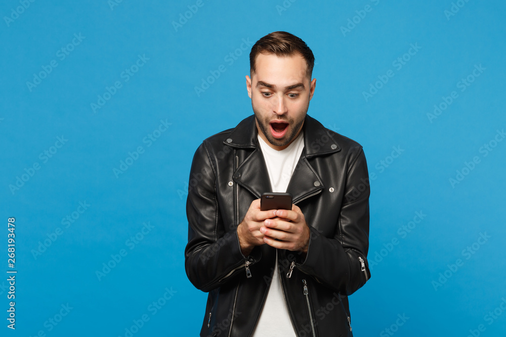 Handsome fun overjoyed excited young unshaven man in black leather jacket white t-shirt using mobile phone isolated on blue wall background studio portrait. People lifestyle concept Mock up copy space Handsome fun overjoyed excited young unshaven man in black leather jacket white t-shirt using mobile phone isolated on blue wall background studio portrait. People lifestyle concept Mock up copy space