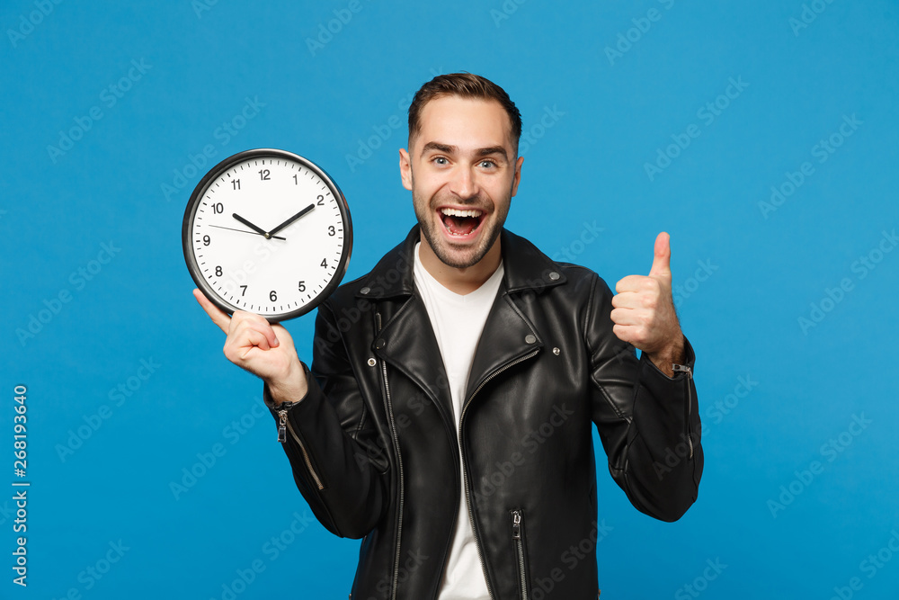 Handsome stylish young unshaven man in black leather jacket white t-shirt holding round clock isolated on blue wall background studio portrait. People lifestyle concept. Hurry up. Mock up copy space. Handsome stylish young unshaven man in black leather jacket white t-shirt holding round clock isolated on blue wall background studio portrait. People lifestyle concept. Hurry up. Mock up copy space.