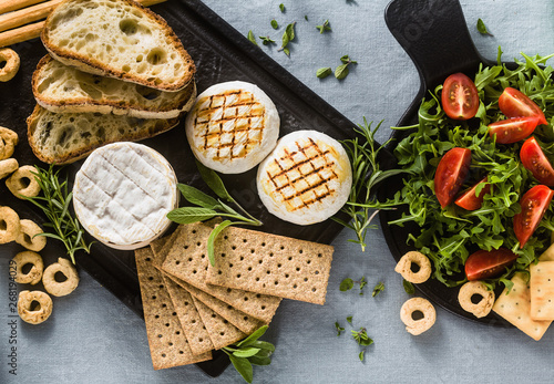 grilled Italian Tomino cheese served on a table with white wine, crackers, grissini and taralli with aromatic herbs and arugula and tomato salad on a blue linen festive tablecloth. summer menu photo