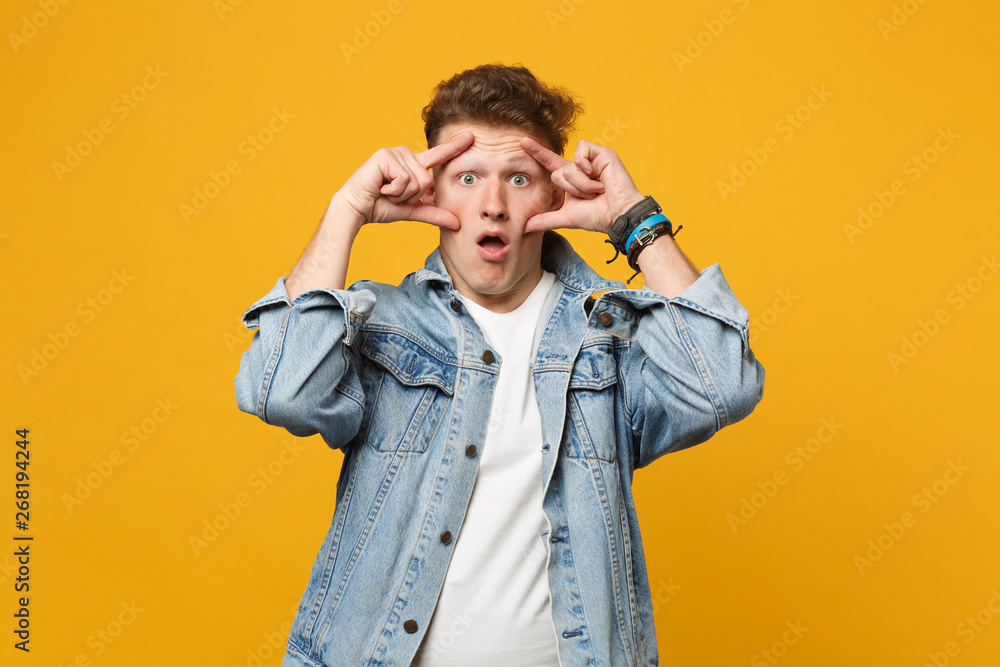 Portrait of shocked young man in denim casual clothes keeping mouth open, stretching eyelids isolated on yellow orange wall background. People sincere emotions, lifestyle concept. Mock up copy space. Portrait of shocked young man in denim casual clothes keeping mouth open, stretching eyelids isolated on yellow orange wall background. People sincere emotions, lifestyle concept. Mock up copy space.
