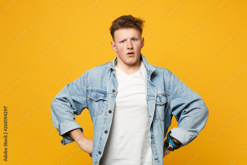 Portrait of shocked perplexed young man in denim casual clothes standing with arms akimbo isolated on yellow orange background in studio. People sincere emotions lifestyle concept. Mock up copy space.
