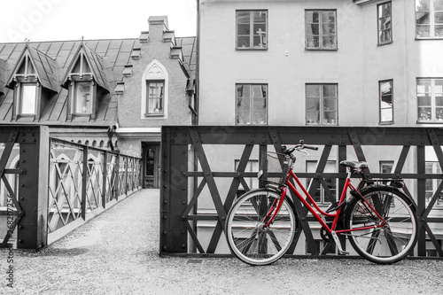 A picture of a lonely red bike standing in the typical street in Stockholm by the bridge to a house. The bike looks to be modern in a retro style. The background is black and white. 