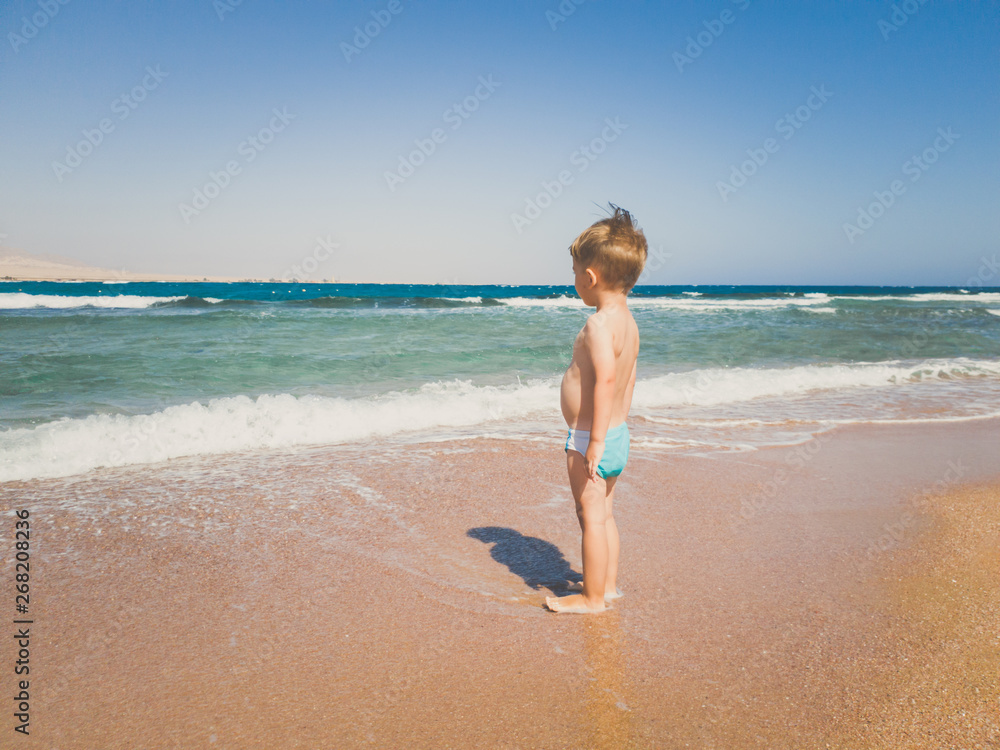 Toned image of 3 years old toddler boy standing on the sea beach and looking at horizon. Child relaxing and having good time during summer holiday vacation.