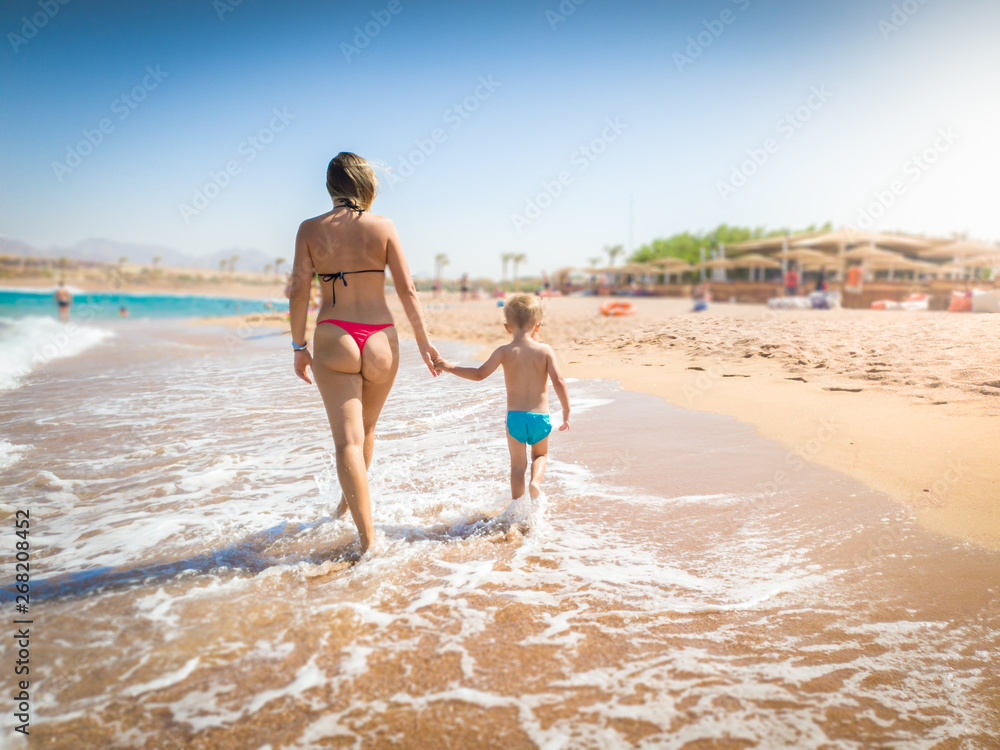 Beautiful young mother holding her little son by hand and walking on the sandy sea beach at bright sunny day. Child relaxing and having good time during summer holiday vacation.