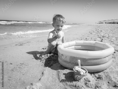 Black and white photo of toddler boy playing on the sea beach with inflatable swimming pool. Child relaxing and having good time during summer holiday vacation.