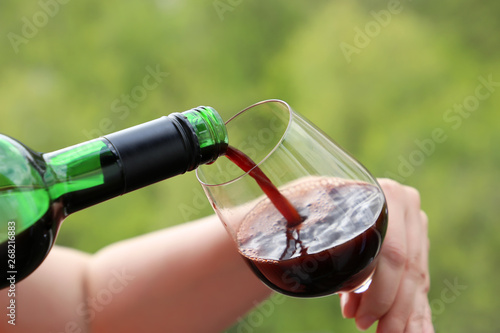 Woman pours red wine from the bottle into the glass on green nature background. Concept of wine drinking outdoors, street restaurant, alcohol tasting at winery