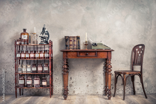 Cuadro en lienzo Old laboratory bottles, vintage microscope, glassware support on the shelving, classic typewriter and antique books on wooden table and chair front concrete wall background
