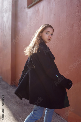 Young beautiful girl with long hair in black coat on sunny day turns around against the red wall. Street style portrait