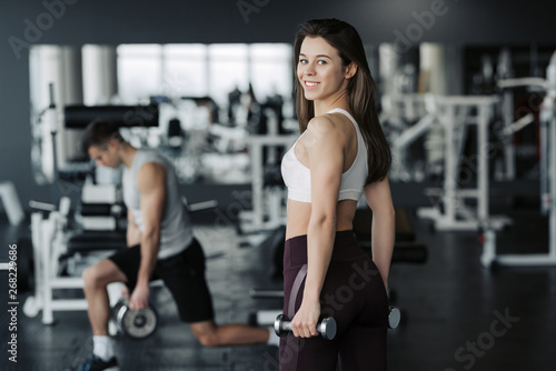Young sportswoman lifting weights in gym wearing sportswear with her boyfriend on background.