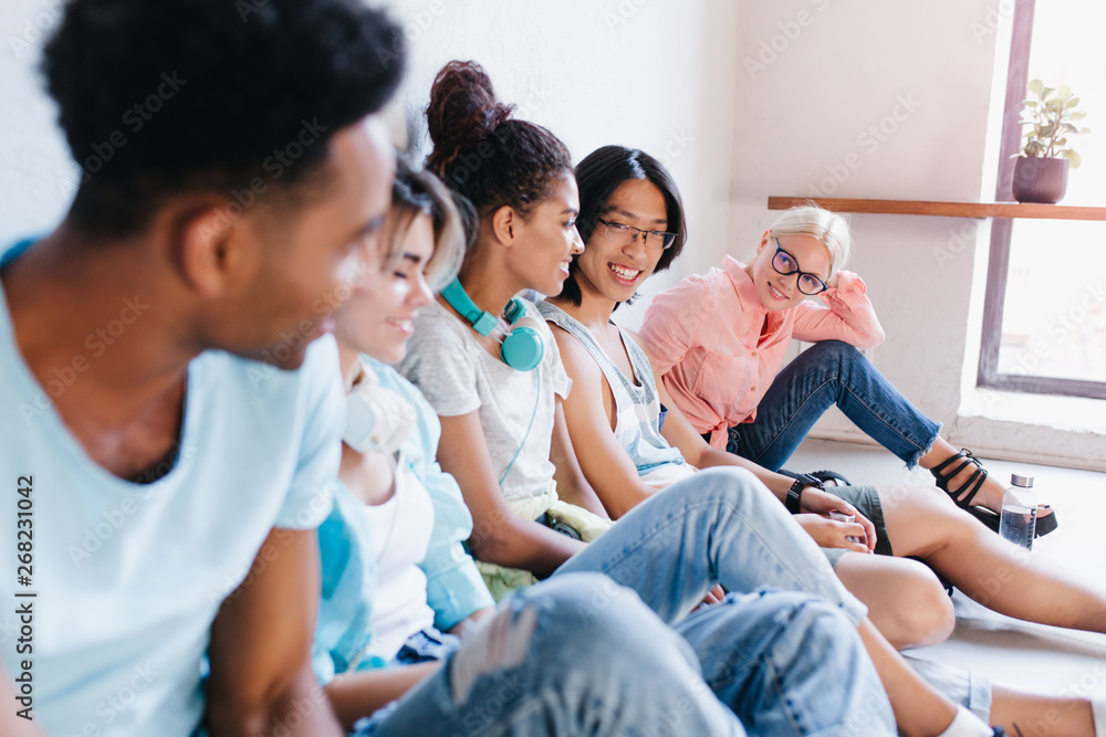 Blonde girl in glasses and pink shirt sitting on the floor and looking with interest at her international classmates. Portrait of students chilling in campus Blonde girl in glasses and pink shirt sitting on the floor and looking with interest at her international classmates. Portrait of students chilling in campus