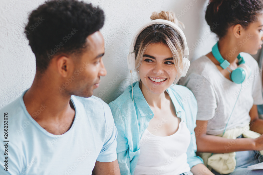 Fototapeta premium Attractive girl in white earphones looking with smile at her african friend in blue shirt. Black young man listening what his charming university mate talking.