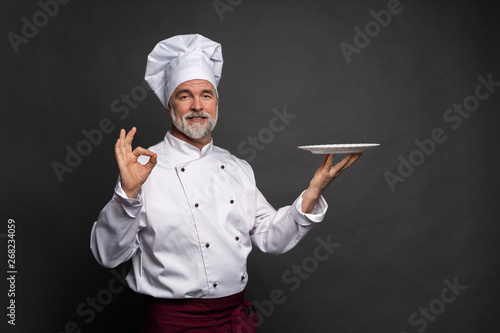 Fotografia Mature cook chef holding an empty plate on a black background.