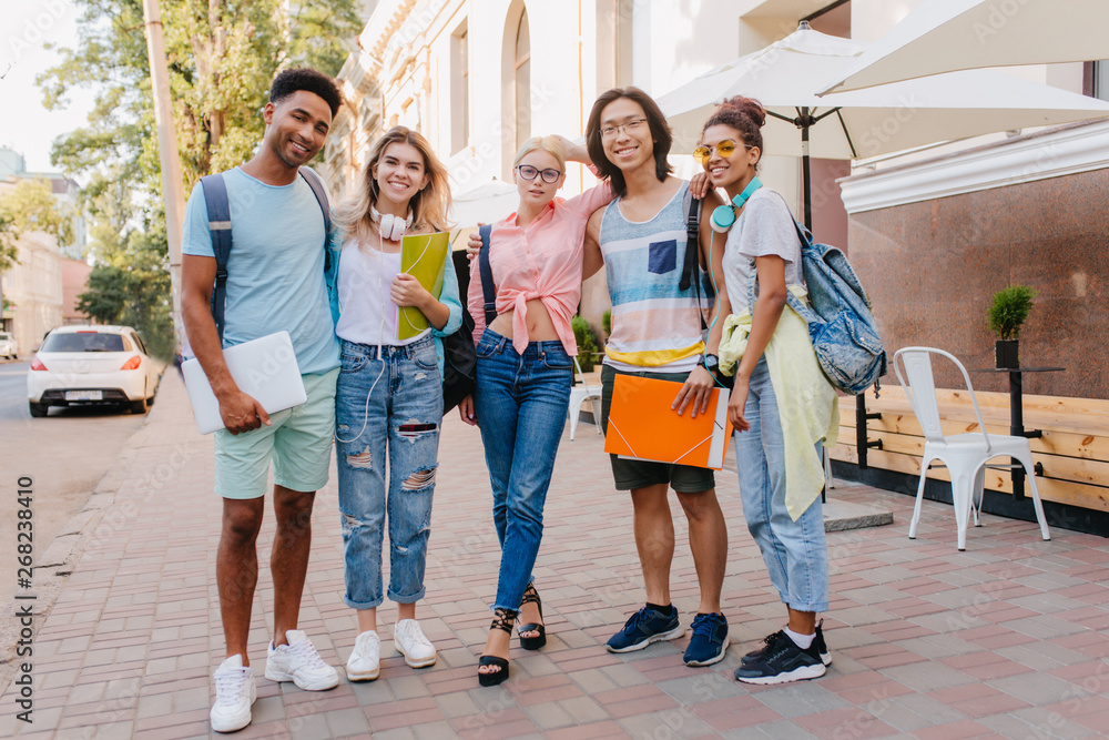 Smiling students with colorful folders and backpacks posing outdoor in morning. Laughing friends of different nationalities standing beside cafe after exams .