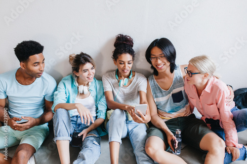 Pretty african girl with blue earphones showing her university mates something interesting in smartphone. International students sitting on floor and listening with interest what their friend tell.
