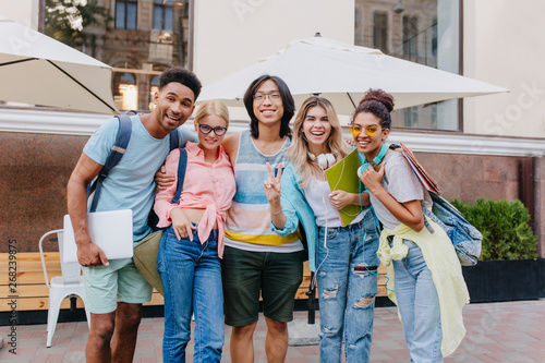 Obraz na plátně Laughing asian boy in glasses and shorts embracing charming blonde girls in front of outdoor cafe